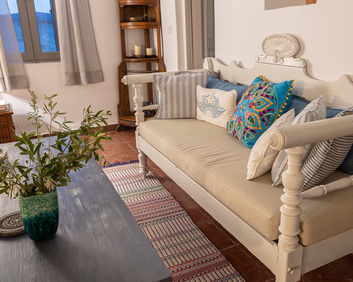 A carved white daybed at Casa di Lavalle piled with striped linen and embroidered Suzani cushions, on terracotta tile beside an antique corner shelf.